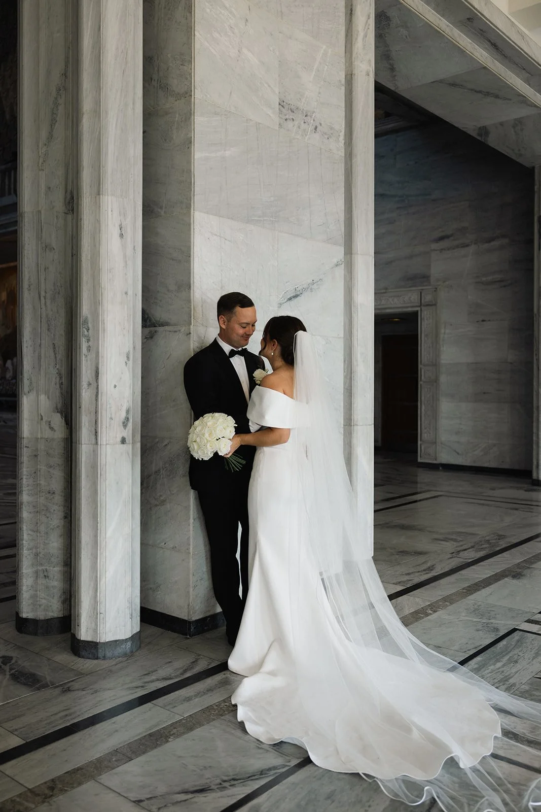 En brud og brudgom står nær hverandre i et marmorgulv og vegg, med brudens lange hvite kjole og slør, og brudgommen i svart tuxedo. De holder en bukett med hvite blomster og ser på hverandre kjærlig.