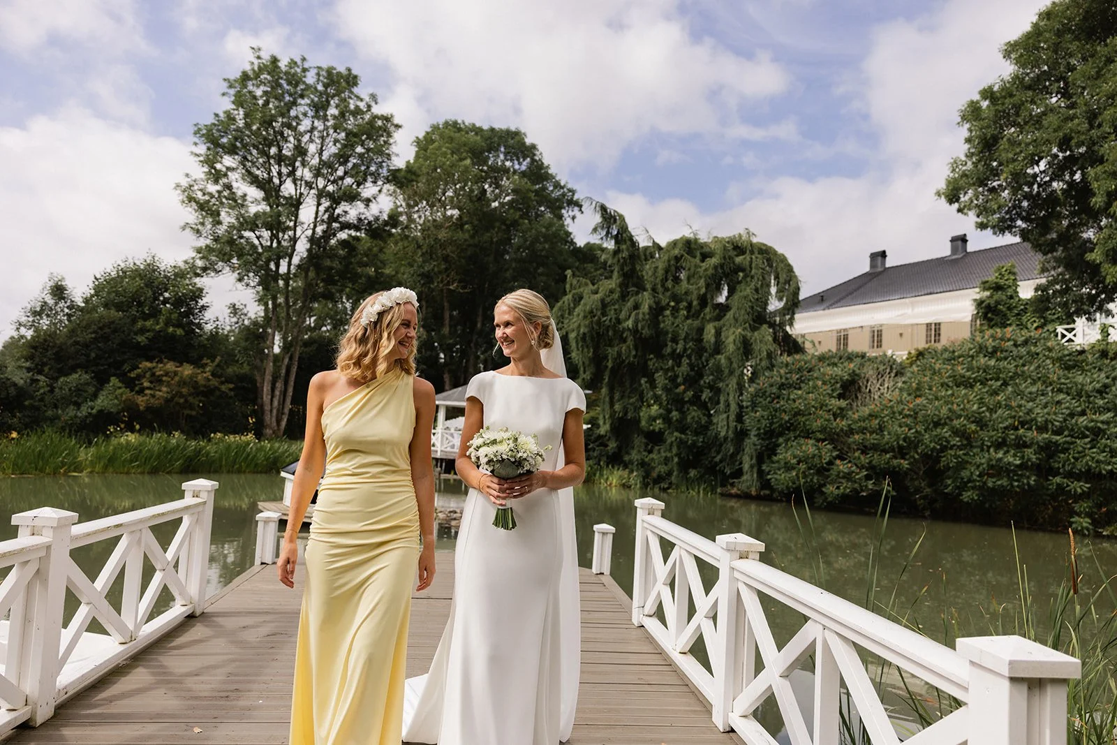 To kvinner, en i en lysegul kjole og en i en hvit brudekjole, går på en trebro over en innsjø, smilende og ser på hverandre, omringet av grønne trær og en bygning i bakgrunnen.