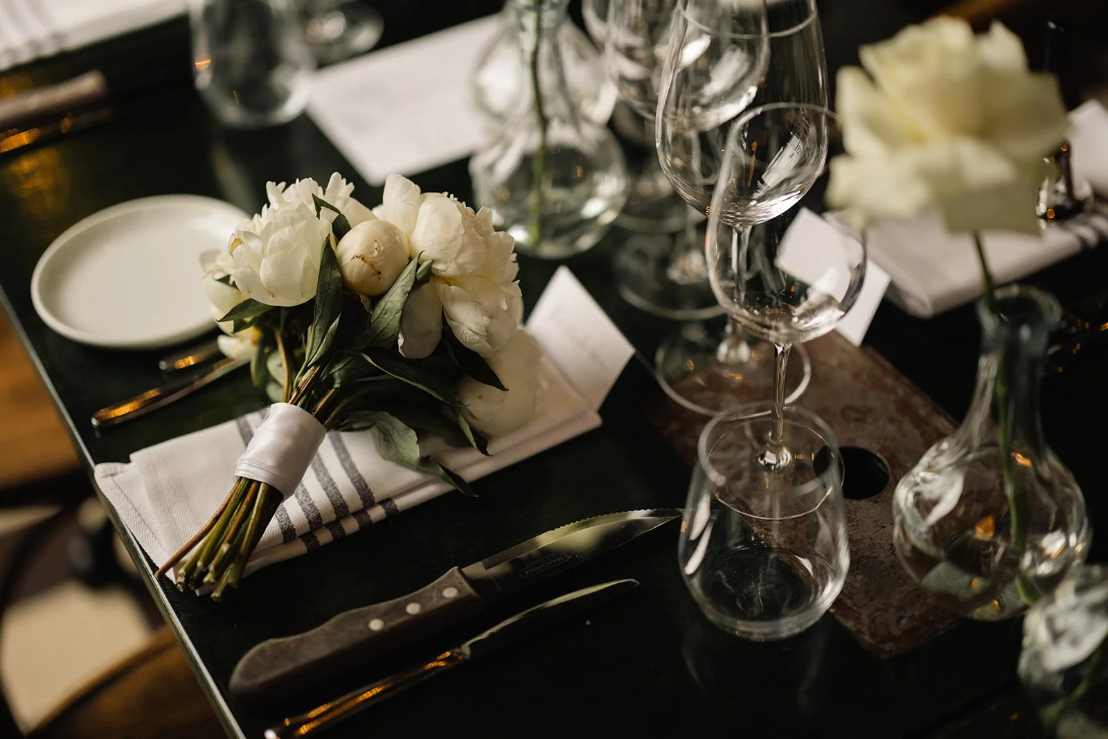 Et bord dekket med hvite blomsterbuketter, tomme vinglass, små hvite kort og en kniv. Bordet er dekket med hvite og svarte tekstiler.