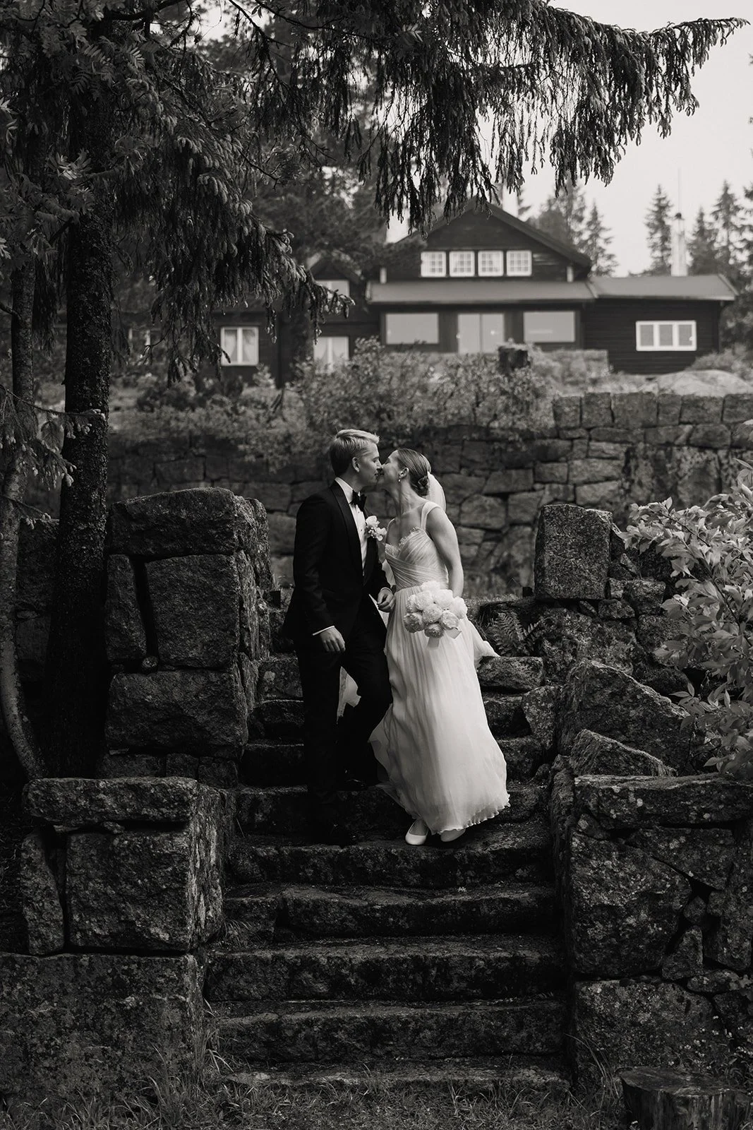 Et svart-hvitt bilde av et bryllup, der en brud og en brudgom kysser på en gammel trapp omgitt av store steiner, trær og en husbakgrunn.