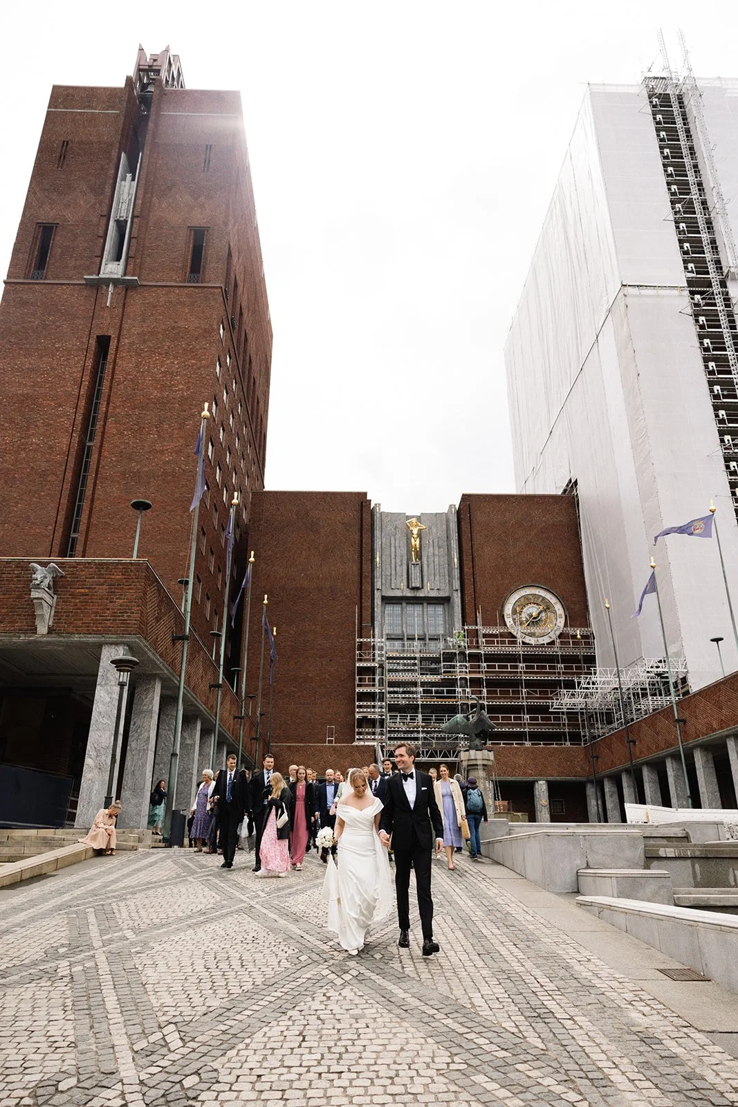 Bryllupsparet går hånd i hånd foran rådhuset, omgitt av gjester, med store bygninger og en klokke i bakgrunnen.