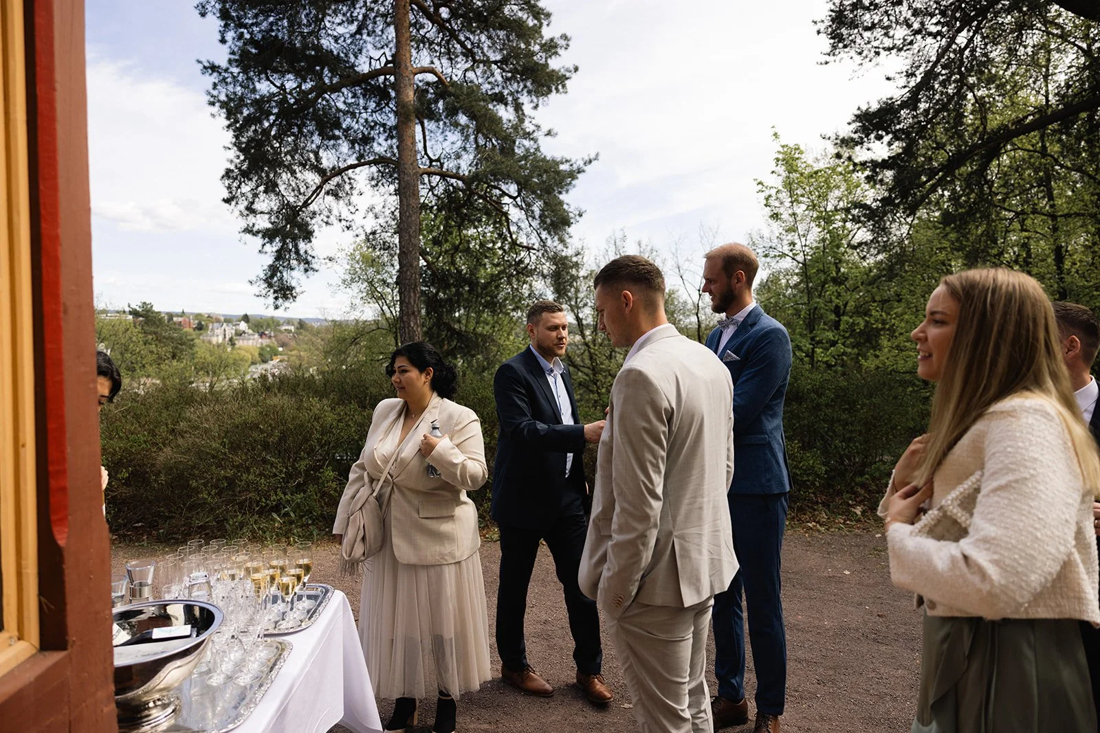 Gruppemennesker kledd i formelle klær, stående ute i en parklignende setting, med et bord med glass og drikke varer.