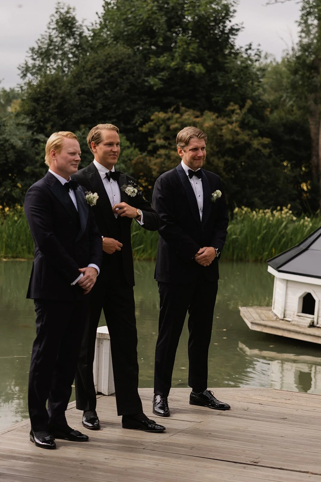 Tre menn i smoking står på en treplattform nær en innsjø, med trær og busker i bakgrunnen.