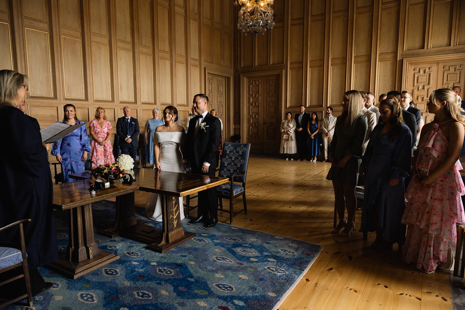 En bryllupsseremoni med brud og brudgom foran en seremoniell offisiant i et rom med trepaneler og lyse trevegger, omgitt av gjester.