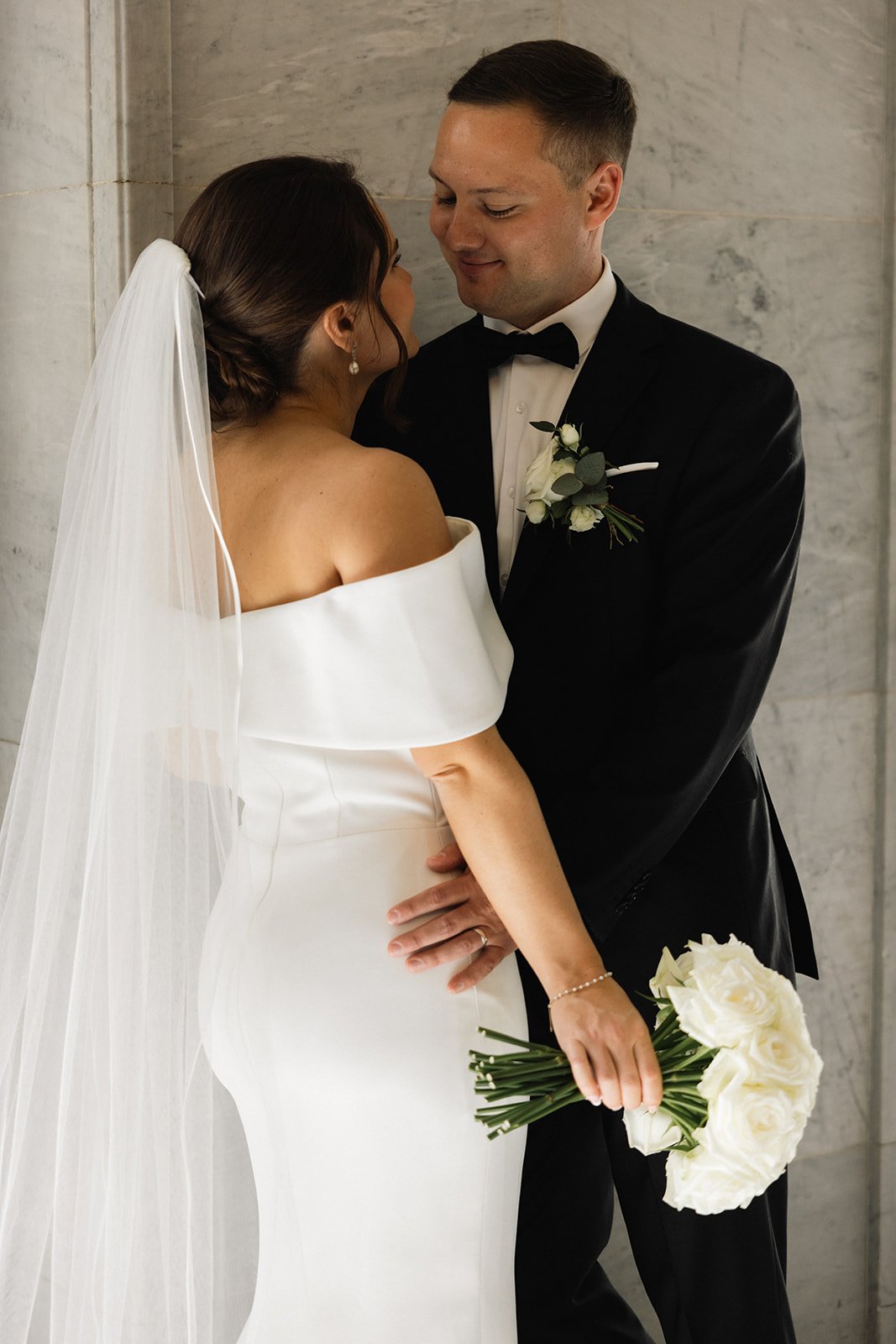 Bryllupspar klemmer, kvinnen har på seg en hvit kjole og brudebånd, mannen er i svart dress og slips, kvinnen holder en bukett hvite roser.