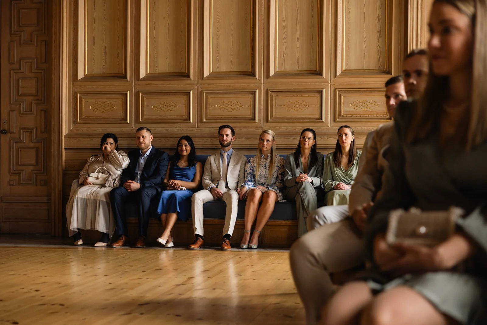En gruppe mennesker sitter på en benk i et rom med treskodd vegg. De er kledd i formelle klær og virker å være i et formelt arrangement.