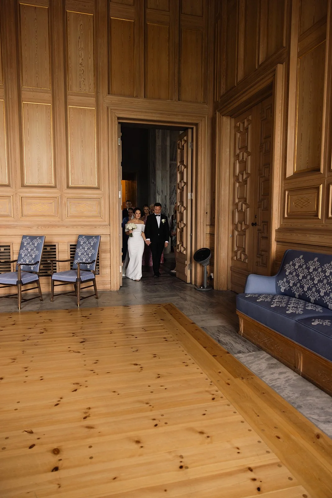 En brud og en brudgom går inn i et rom med trepanelvegger under en seremoni, omgitt av gjester.