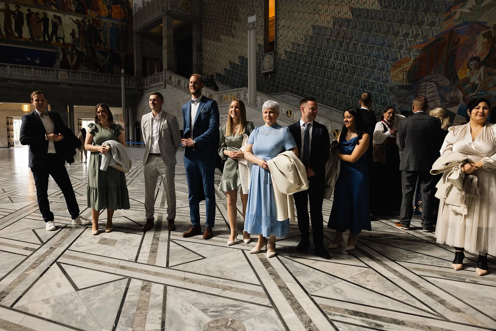 En gruppe mennesker kledd i formelle klær står i en stor hall med marmorgulv og et fargerikt veggmaleri i bakgrunnen.