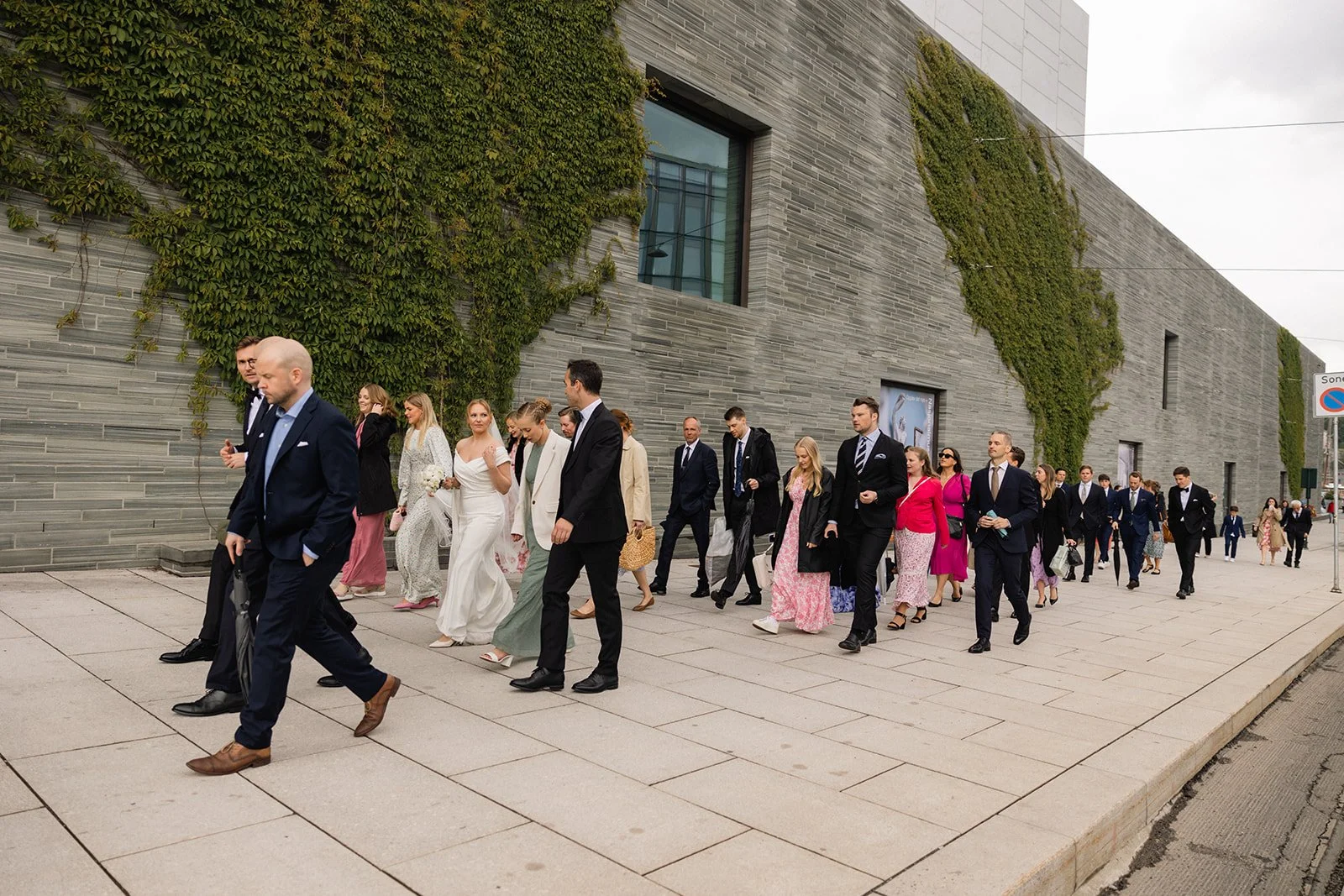 En gruppe mennesker kledd i formell antrekk går langs en bygate utenfor en bygning som har vegger dekket med grønn vegetasjon.