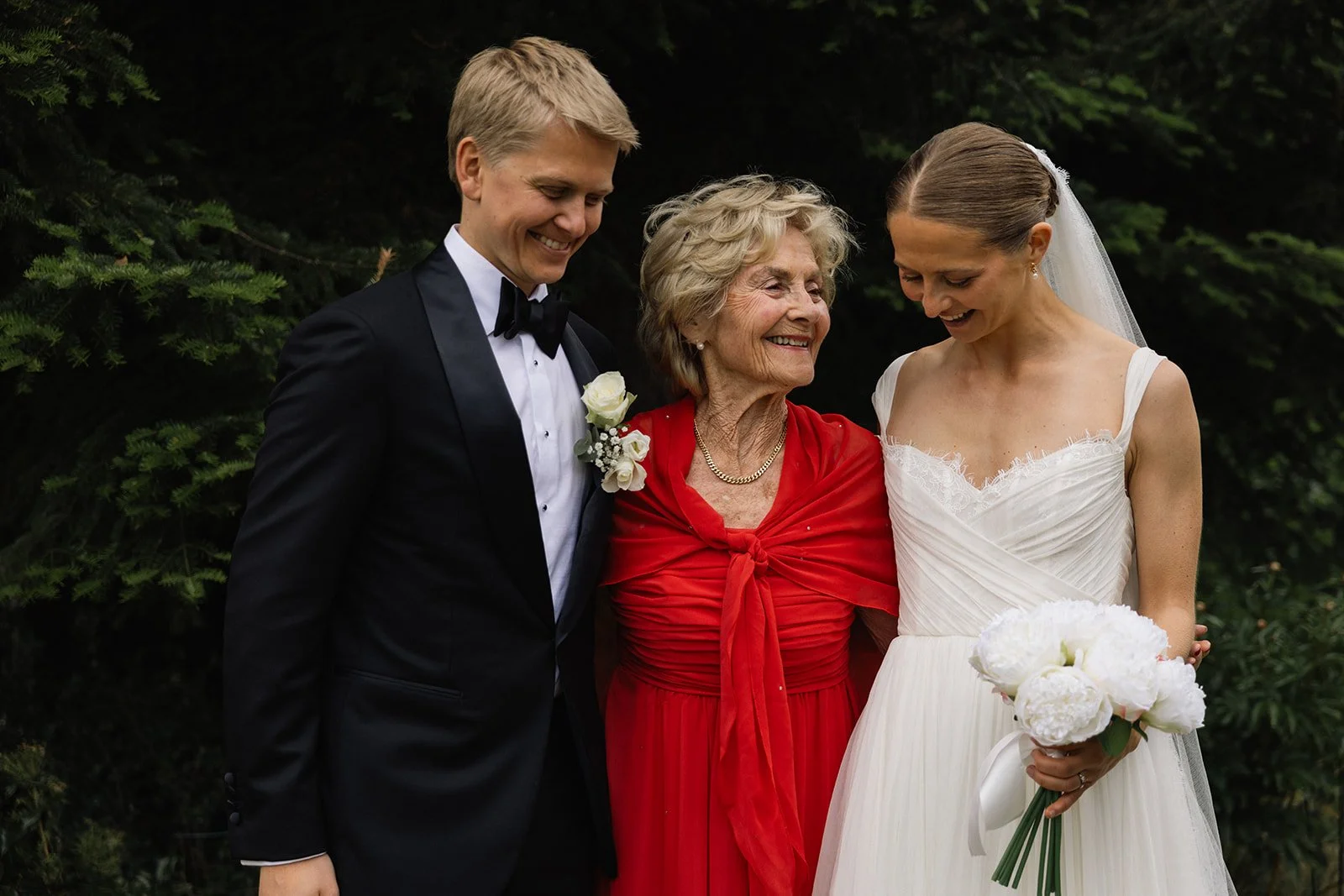 Et bryllupsbilde av en ung brud og en ung brudgom som smiler og står sammen med en eldre kvinne som antagelig er moren hennes. De er ute i naturen med grønne trær i bakgrunnen.