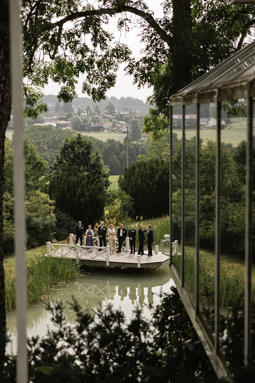 Et bryllupsselskap står på en liten, hvit treplattform i en innsjø, omgitt av grønt landskap og trær, sett fra et vindu.