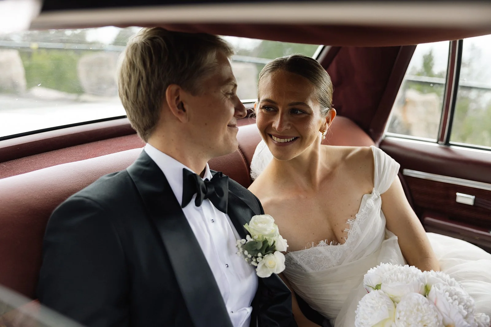 En brudgom og en brud sitter sammen i en bil, smiler og har øyekontakt. Bruden holder en bukett med hvite blomster.