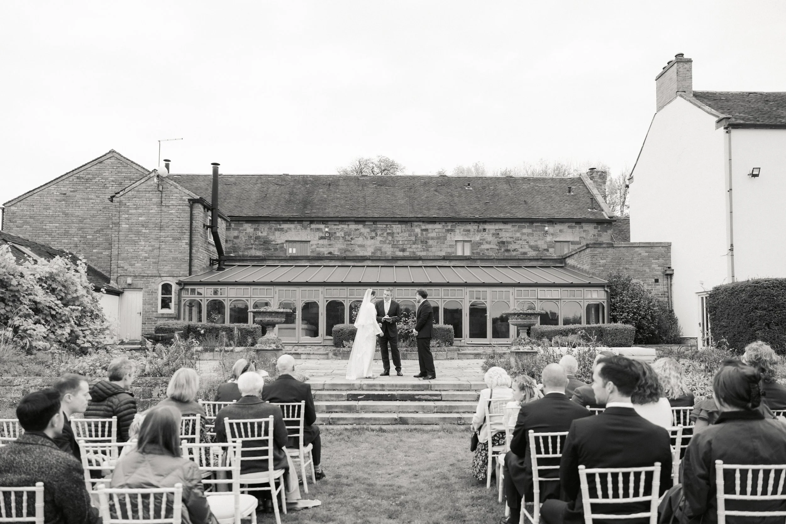 Et utendørs bryllup med gjester sittende på hvite stoler, ser på som et par blir viet av en person inntil en bygning med glassvinduer.