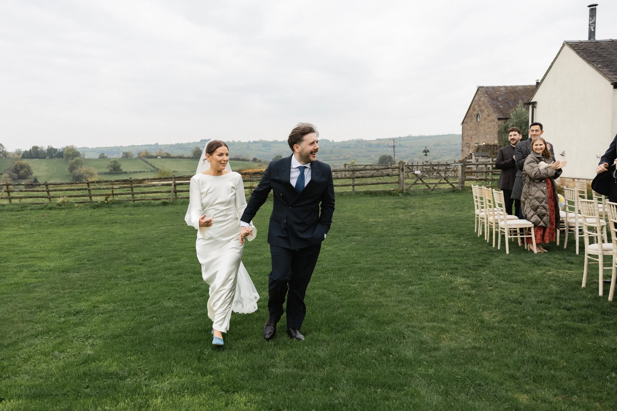 Bride and groom walking across a grassy outdoor area at a wedding reception, with guests seated at tables nearby.