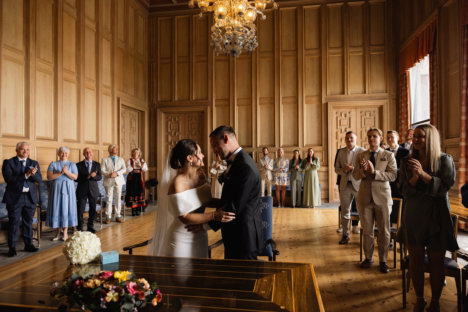 Et brudepar står foran i en seremoni i et rom med trekt trekledning, med venner og familie som applauderer og smiler, mens de holder hverandre i hendene.