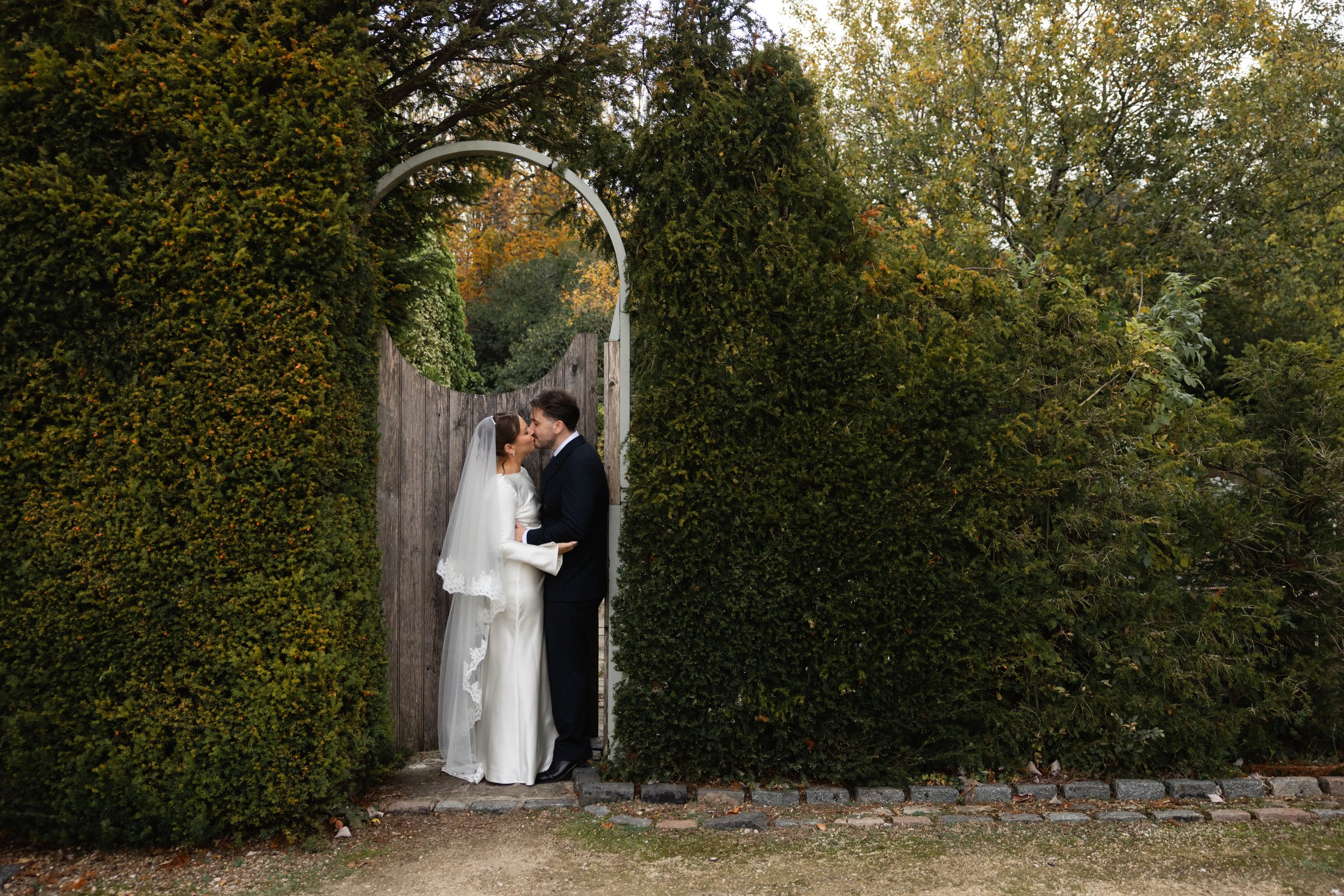 Et nygift par står under en halvmåneformet port omgitt av grønne busker og trær i høstfarger.