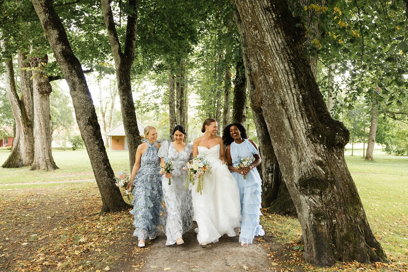 En gruppe kvinner i bryllapskjole og pastellfargede kjoler går gjennom en park med store trær, alle smiler og bærer blomsterbuketter.
