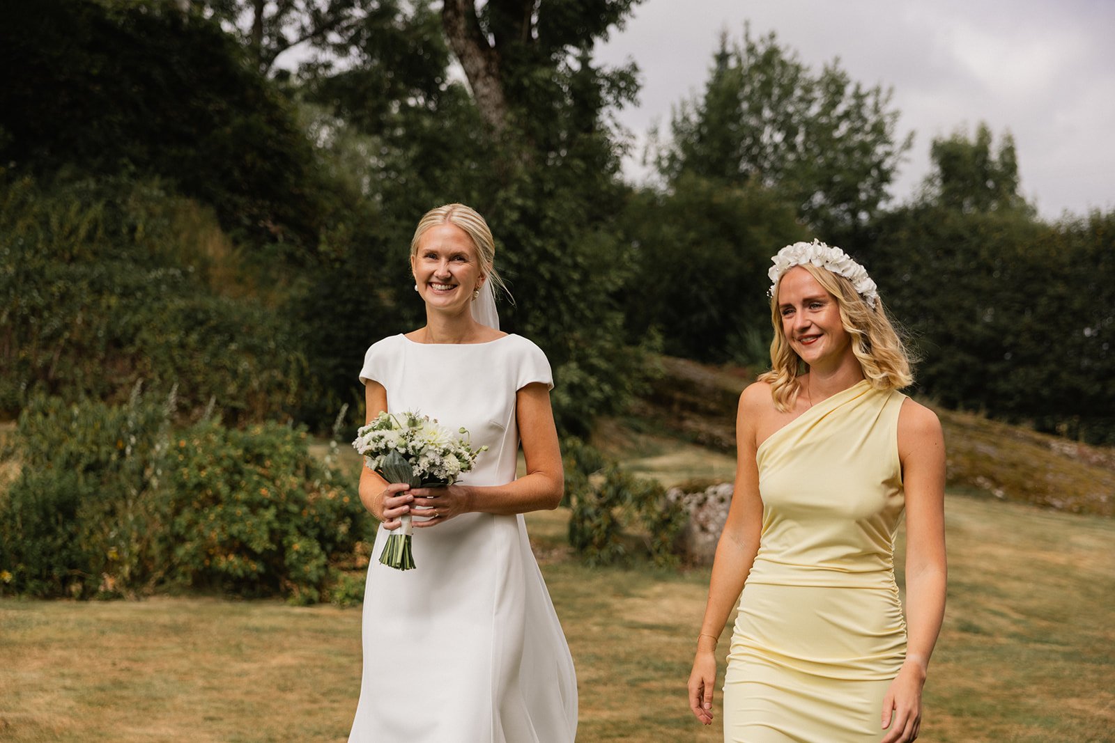 To kvinner går utendørs på en grønn eng, en av dem er i en hvit brudekjole som holder en bukett blomster, den andre er i en lys gul kjole med blomsterkrans i håret.