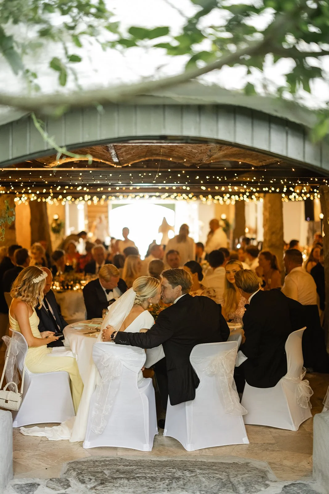 Bryllupsmiddag med mange gjester, der en brud og en brudgom kysser, omgitt av festdeltakere i et innendørsområde dekorert med lys og grønne planter.