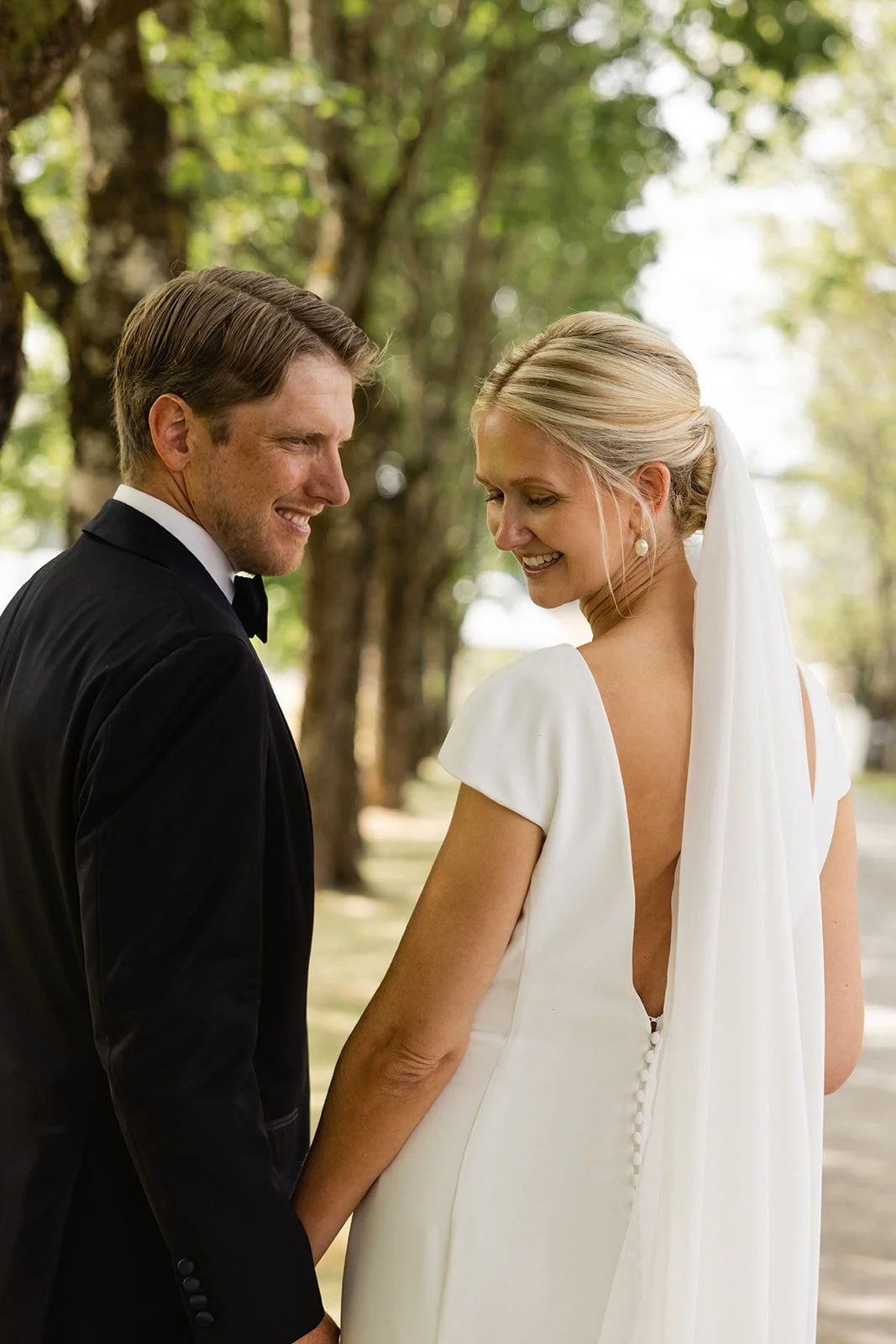 Et brudepar står utendørs under trær, ser på hverandre og smiler. Kvinnen har langt blondt hår, iført en hvit bryllupskjole med åpen rygg og skjult knapper, og har store perleøreringer. Mannen har kort, stylet hår og er iført svart smoking med hvit s