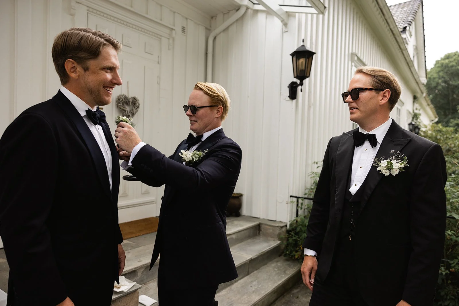 Tre menn i svart dress og slips, to av dem med blomster på venstre bryst, står foran en hvit trapp utenfor en bygning. Den ene mannen blir pyntet med en hvit blomst av en annen mann. Alle har solbriller.
