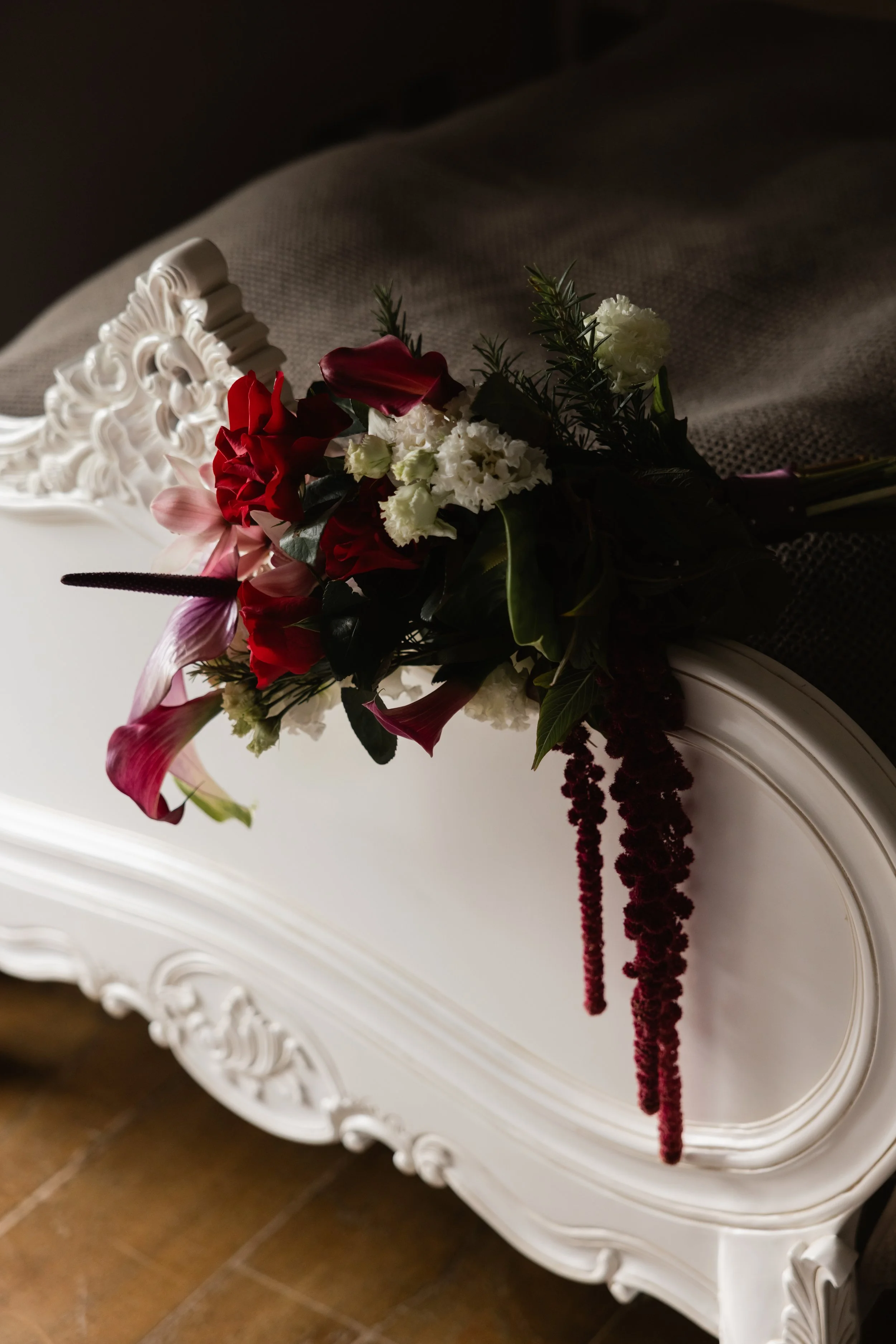 En bukett med blomster plassert på en hvit, utskåret trebord. Buketten inneholder røde, hvite og lillafargede blomster med grønne blader, og noe som ser ut som lange, tette, røde blomsterstilk.