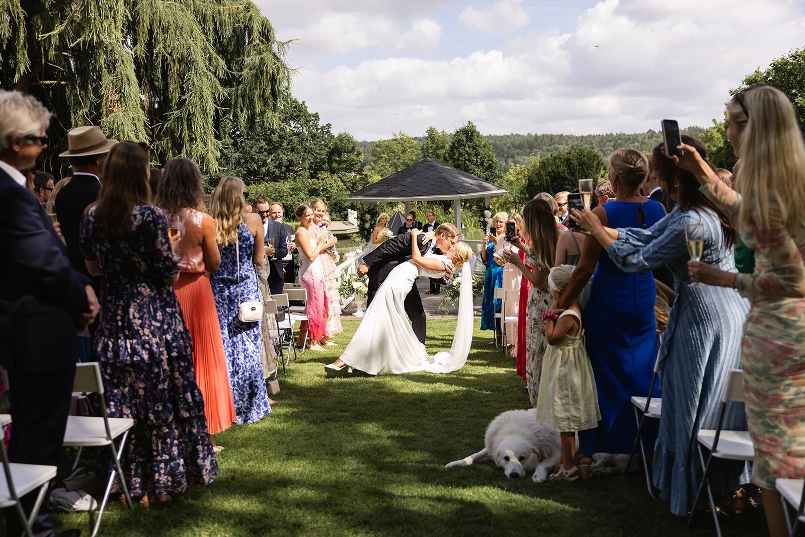 Et bryllup utendørs med mange gjester som ser på en bukett som blir delt ut. En brud og en brudgom kysser, mens en stor hvit hund ligger på bakken.