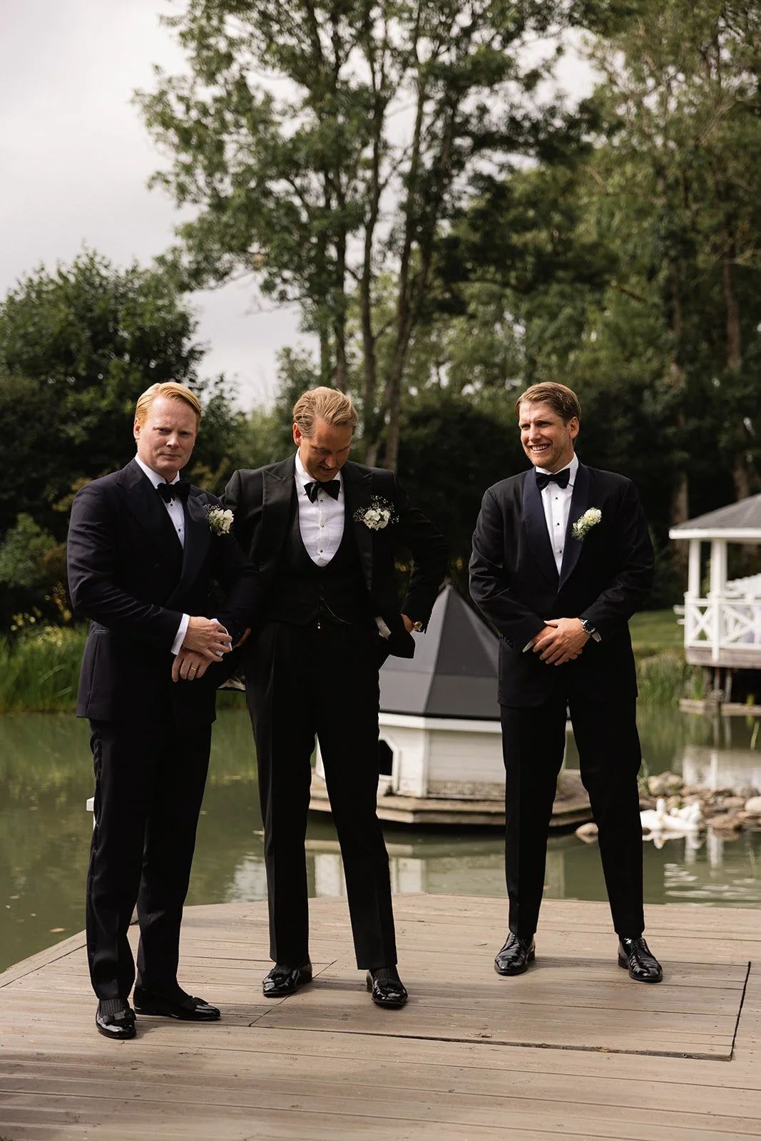 Tre menn i formelle tuxedoer står på en brygge ved en innsjø, smiler og ler, omgitt av trær og en liten hvit brygge i bakgrunnen.