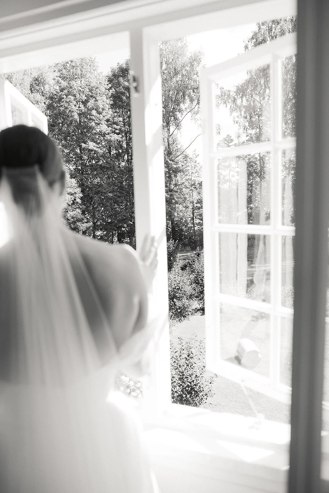 En kvinne med brunt hår i en hvit kjole ser ut av et vindu, med en hage og trær utenfor, i svart-hvitt.