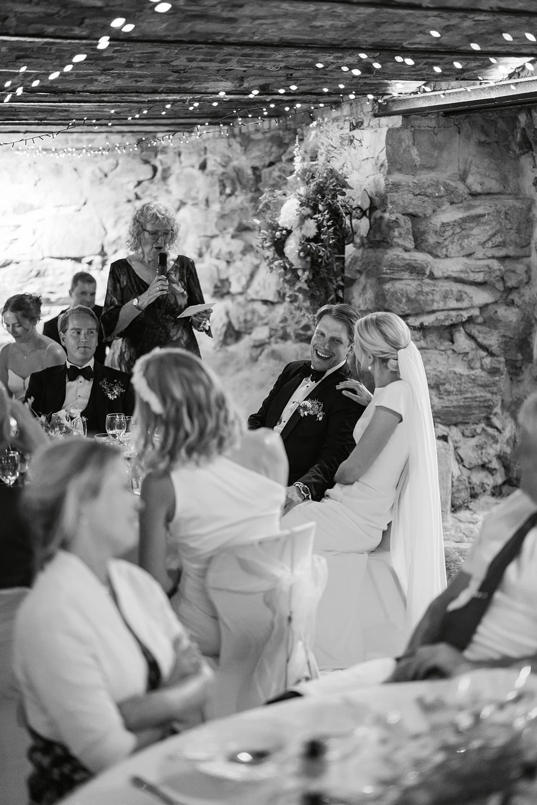 Et bryllupsselskap sitter rundt bord, bruden kikker på mannen sin, som ler. En kvinne står og leser et brev eller tale. Noen personer har på seg formelle klær, og det er dekorasjoner og lys over det rustikke steinrommet.