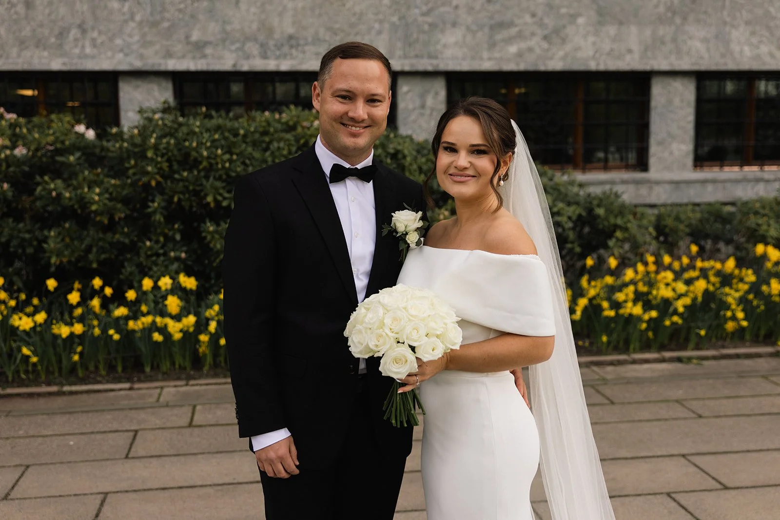 Et nybryllupsbilde av et brystpar i formelle klær, med blomster og smil, foran en blomsterbed og en bygning.