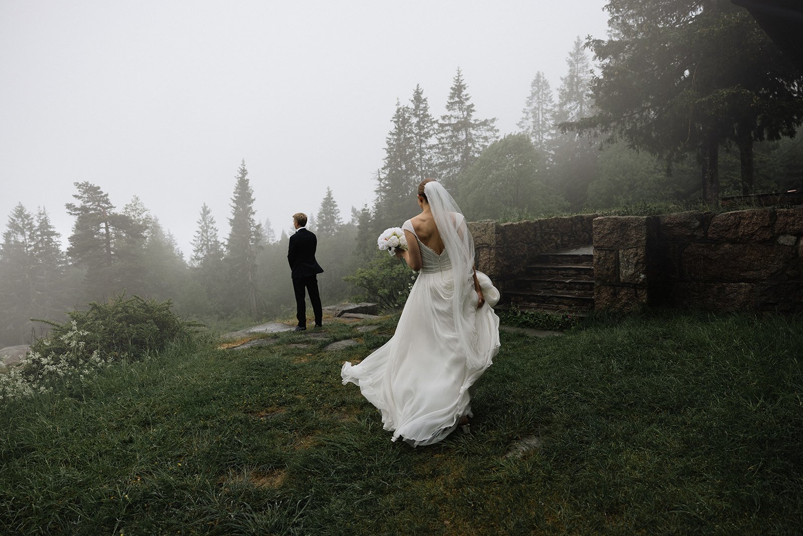 Et brudepar står i en skogbelagt, tåket utendørs setting. Bruden er i en hvit bryllupskjole med lang, flytende kjole og holder en bukett. Brudgommen er i en svart dress og står med ryggen vendt mot kamera.
