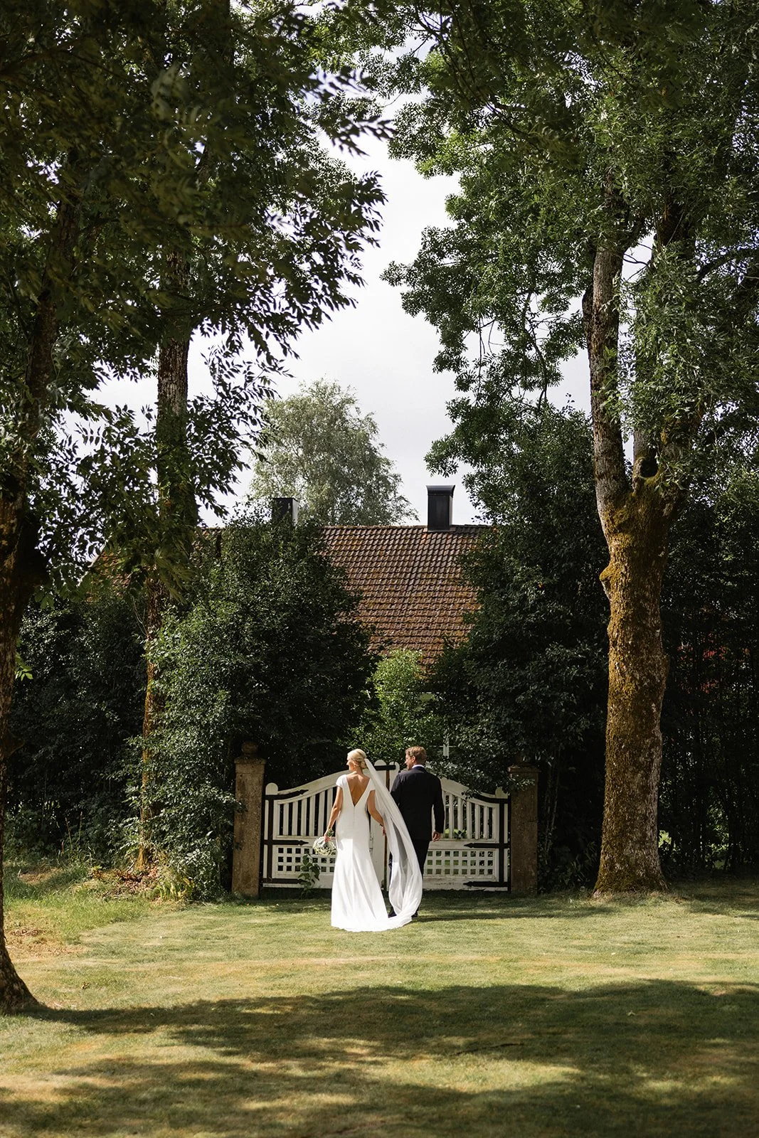 To personer i bryllupsantrekk går mot en hageport, omgitt av store trær.