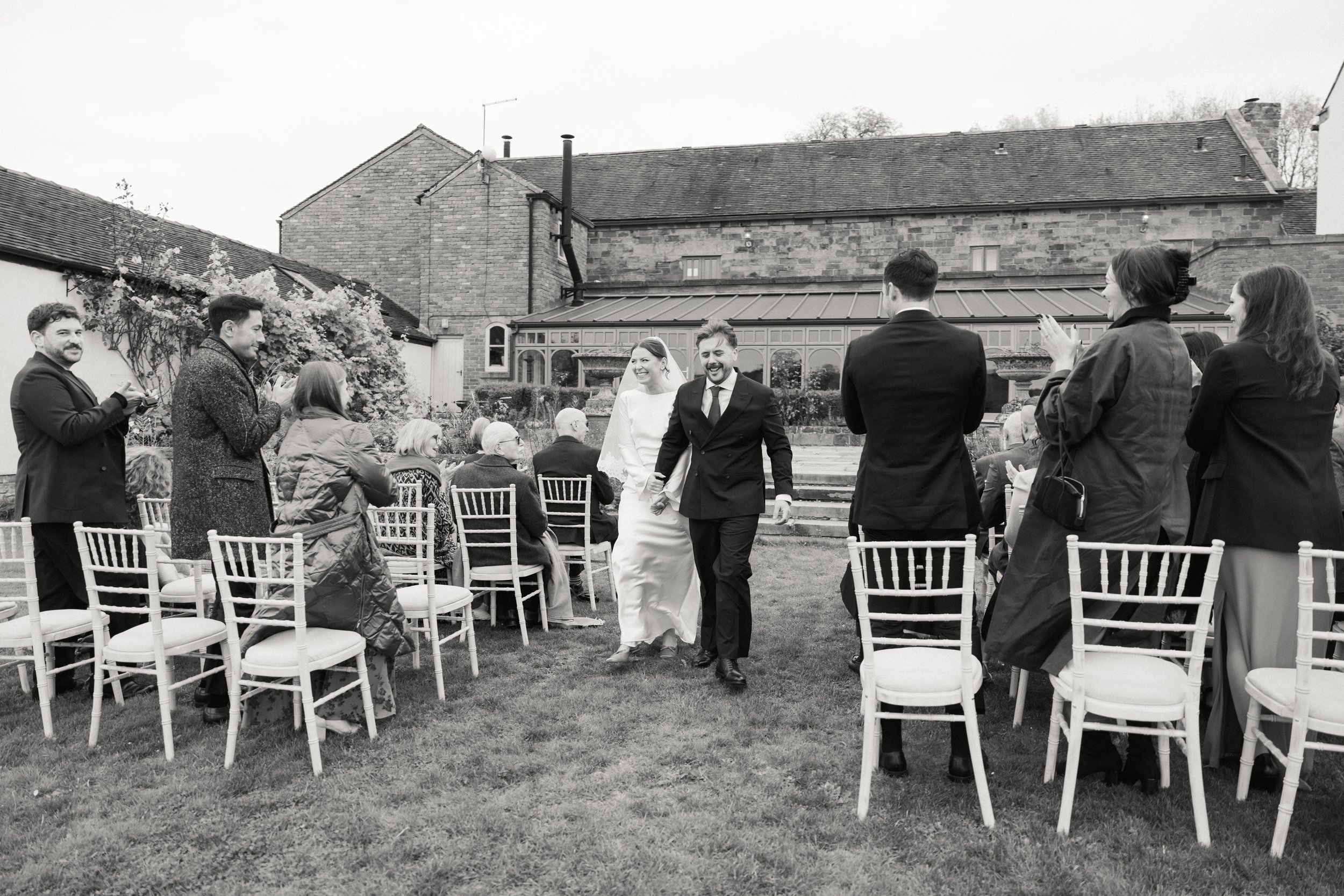 Et svart-hvitt bilde av et bryllup med bruden og brudgommen som går nedover en grastue forbi gjester som applauderer og tar bilder utendørs foran en gammeldags bygning.