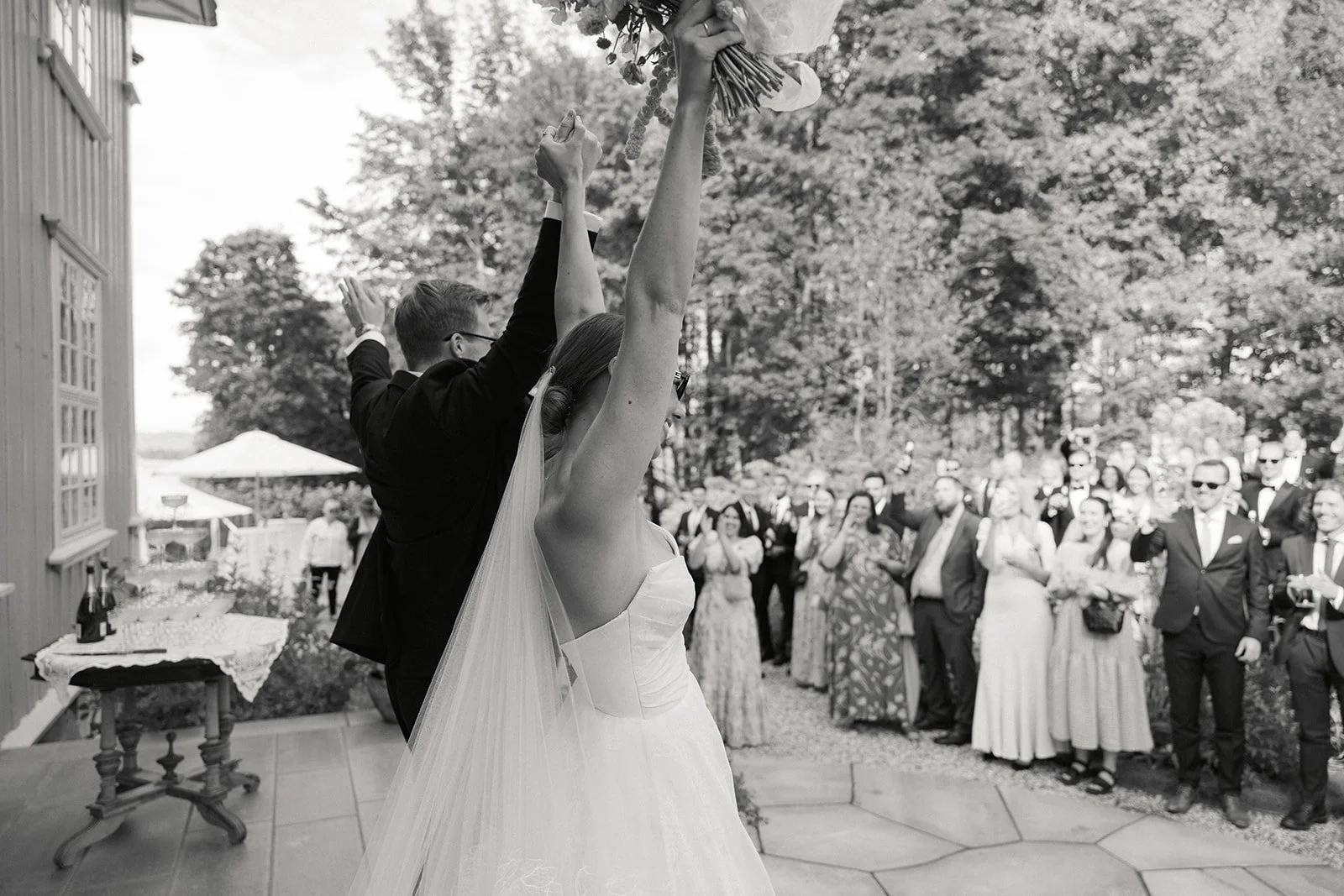 Et vielsepar med brud og brudgom som holder hendene sine opp, mens gjestene ser på og applaudere utendørs.