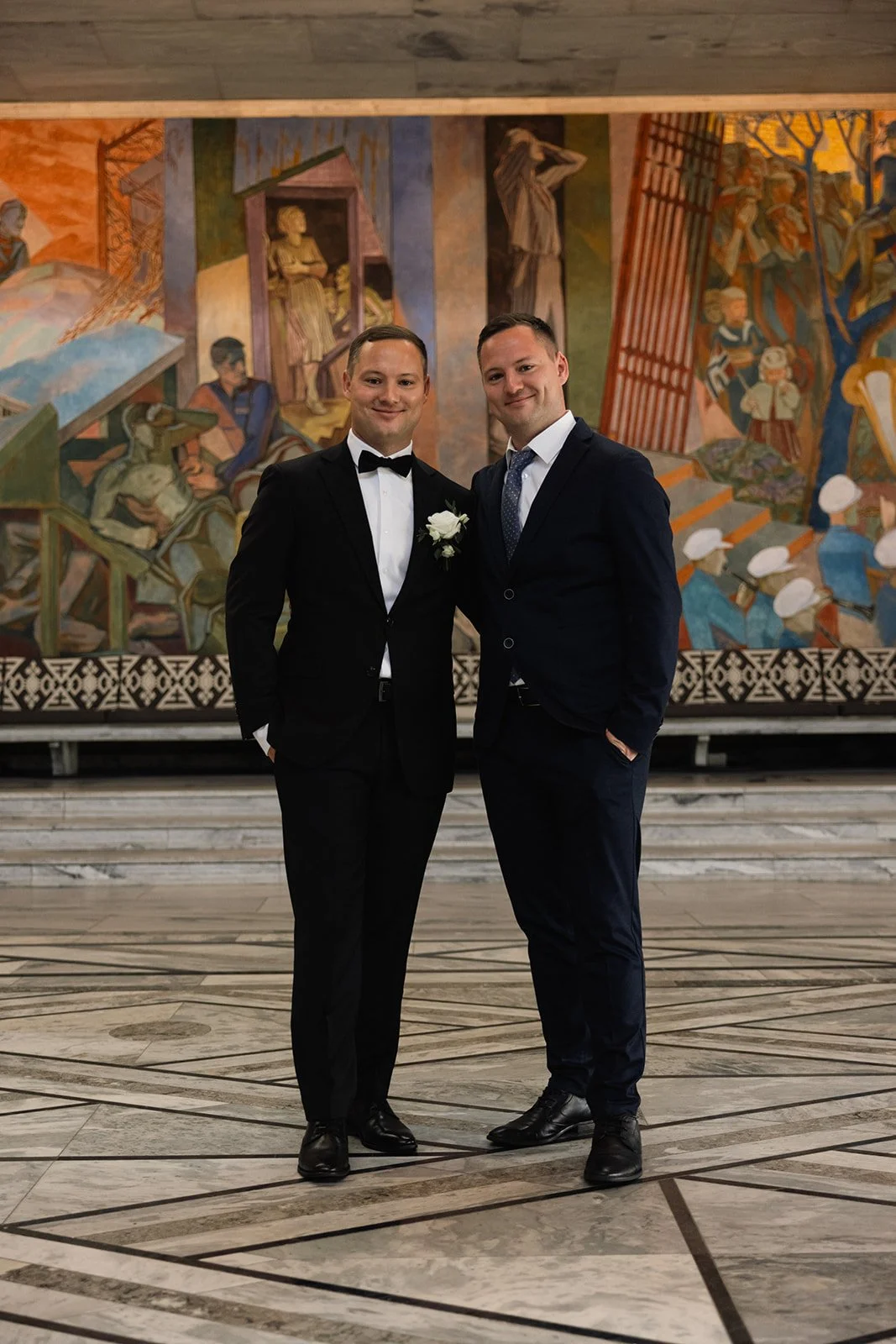 To menn i formelle klær, en i smoking med hvit skjorte og svart sløyfe, og den andre i mørk dress med hvit skjorte og blå slips, står side om side foran en fargerik vegg med malerier.