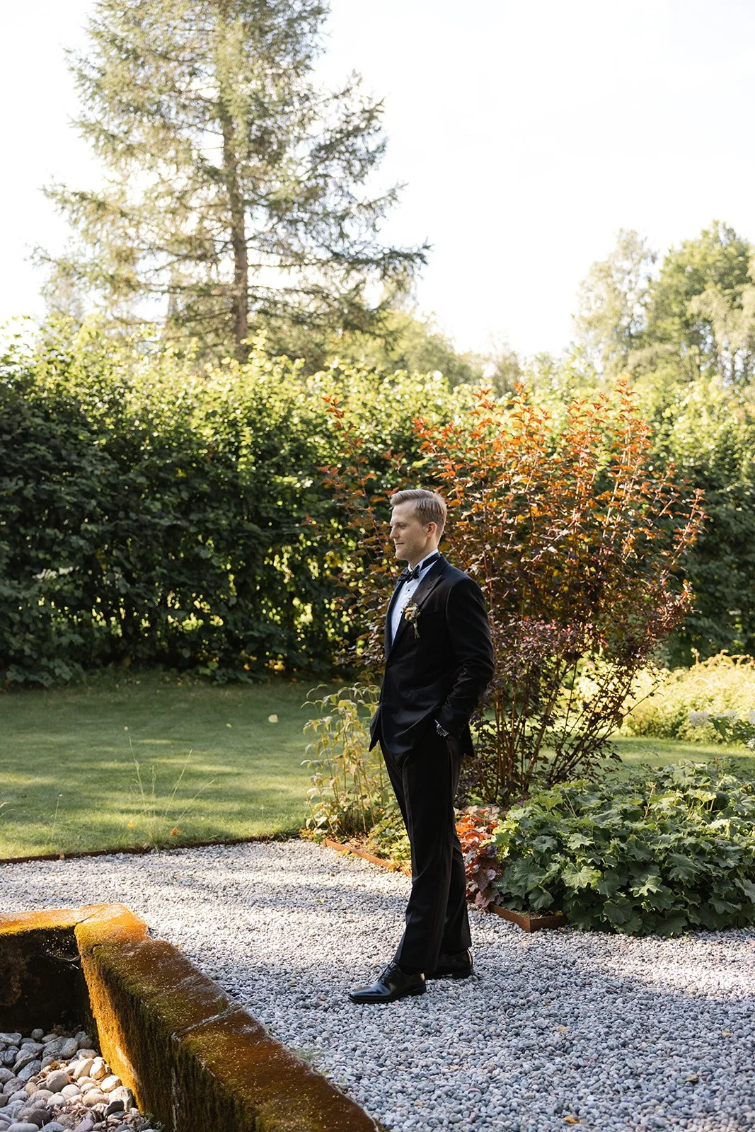 En mann i svart dress og slips, står i en hage med grønne busker og trær, under et klart solrikt himmel, ser forsiktig til høyre.