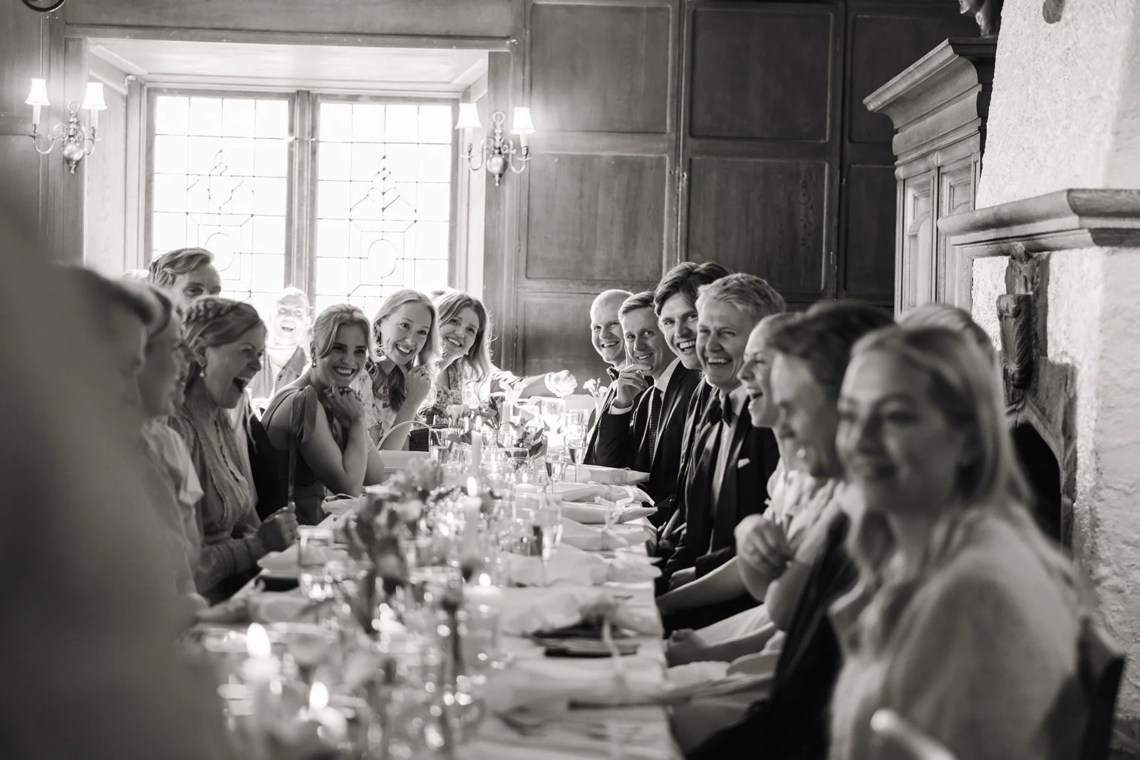 Mennesker som sitter rundt et langbord under en fest i et rom med store vinduer og wood paneling, alle smilende og kledd i formelle klær.
