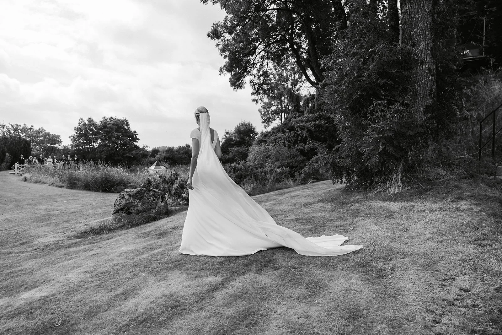 En kvinne i en hvit brudekjole står i en hage med store trær og gress, sett fra ryggen.