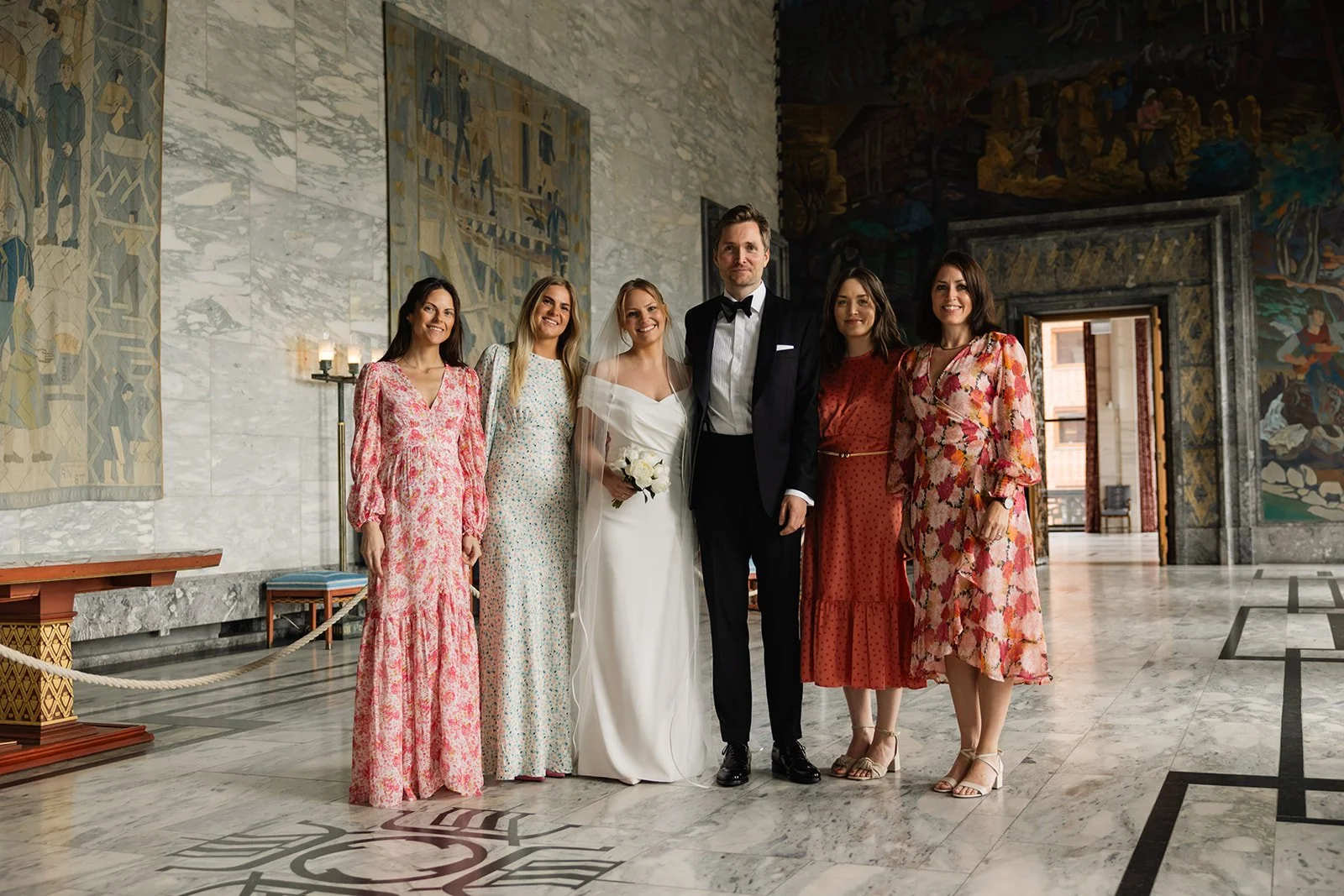 Et bredt gruppemed mennesker på et elegant, kunstnerisk interiør med veggmalerier og marmorgulv, inkludert brud og brudgom sammen med fem kvinner i blomstrede kjoler, sannsynligvis på en bryllupsfeiring.