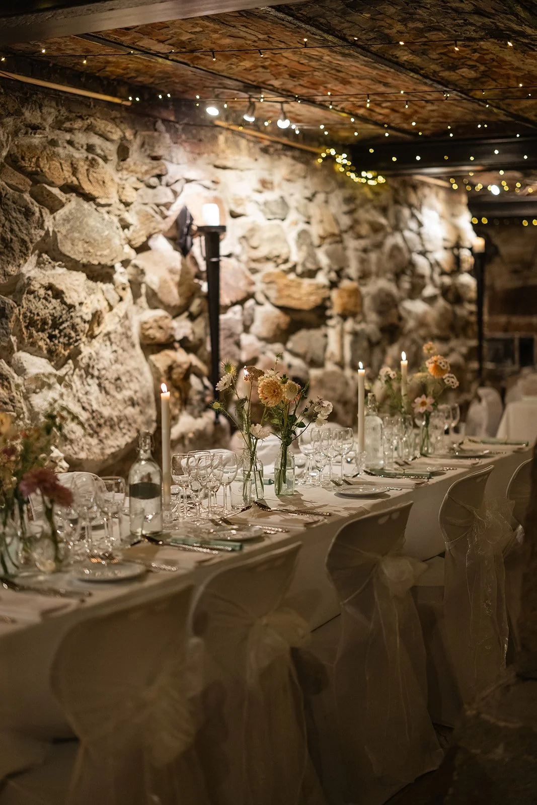 Et lang bord dekket med glass, tallerkener, bestikk og lys i en rustikk restaurant med steinvegger og trehimling med små, varme lys. Dekorative blomsterarrangementer i vaser pynter bordet.