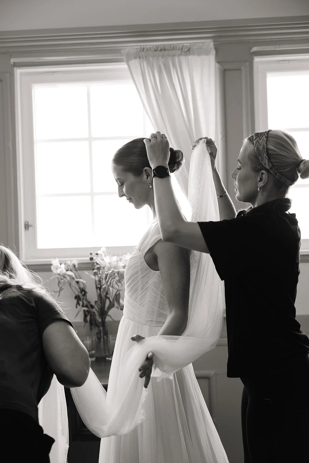 To kvinner hjelper en kvinne med brudekjolen, inntil et vindu og blomster er synlige i bakgrunnen.