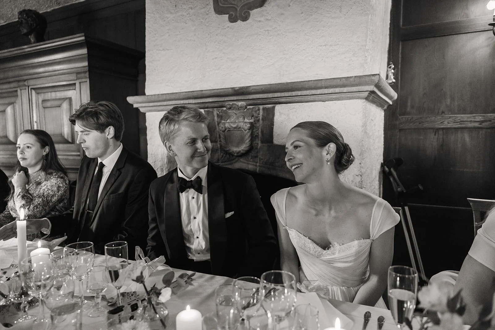Brudeparet med tre andre personer som sitter ved et bord dekket med glass, lys og blomster under en seremoni eller fest.