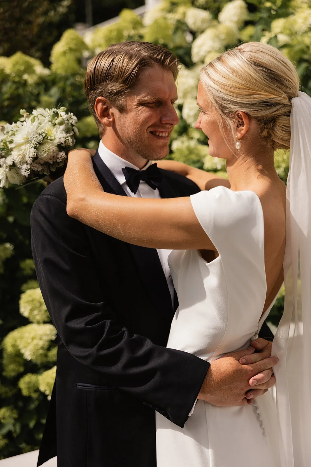 Et brudepar som smiler og omslutter hverandre med hender, foran blomstrende busker.