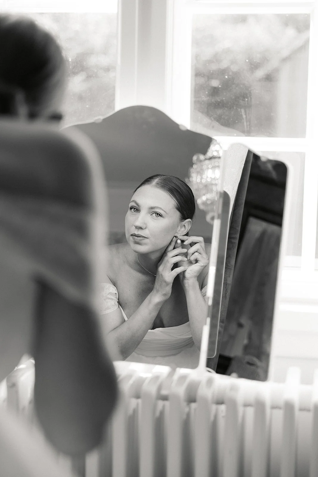 En kvinne ser på seg selv i et speil mens hun setter på øredobber, i svart-hvitt.