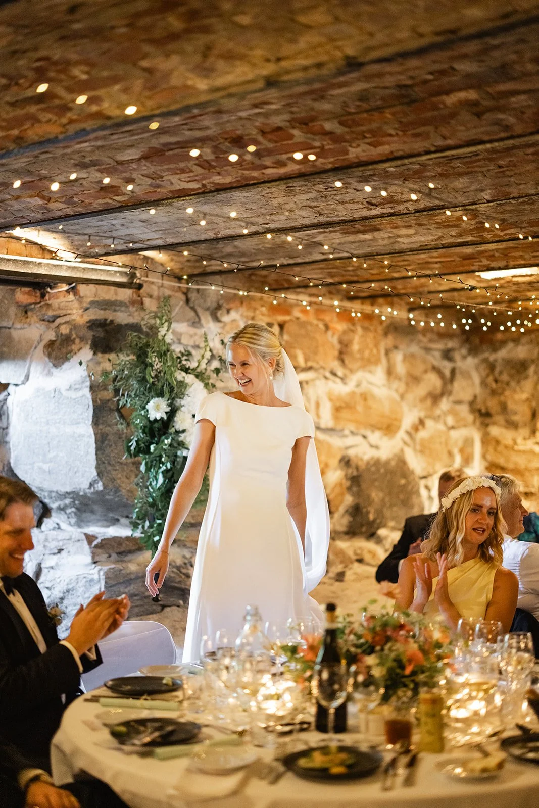 En lykkelig brud i hvit bryllupsklær smiler på bryllupsfest i en rustikk kjeller med mursteinsvegg, dekorert med små lys og blomster.