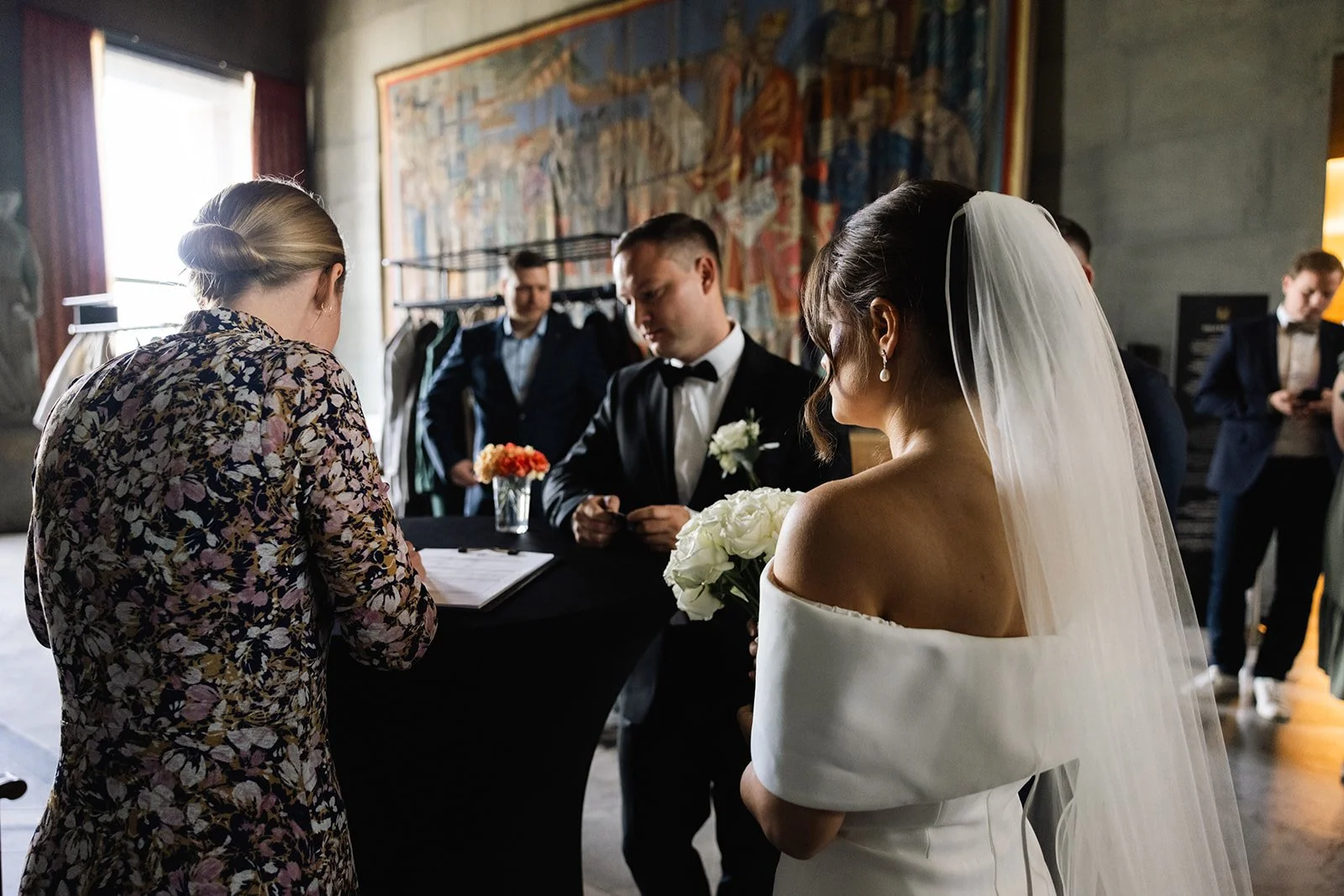 Brude og brudgom står foran en person som leser et bryllupsløfte, med bruden i en hvit kjole og veivende slør, og alle i formelle klær i et rom med kunst på veggen og blomsterdekorationer.