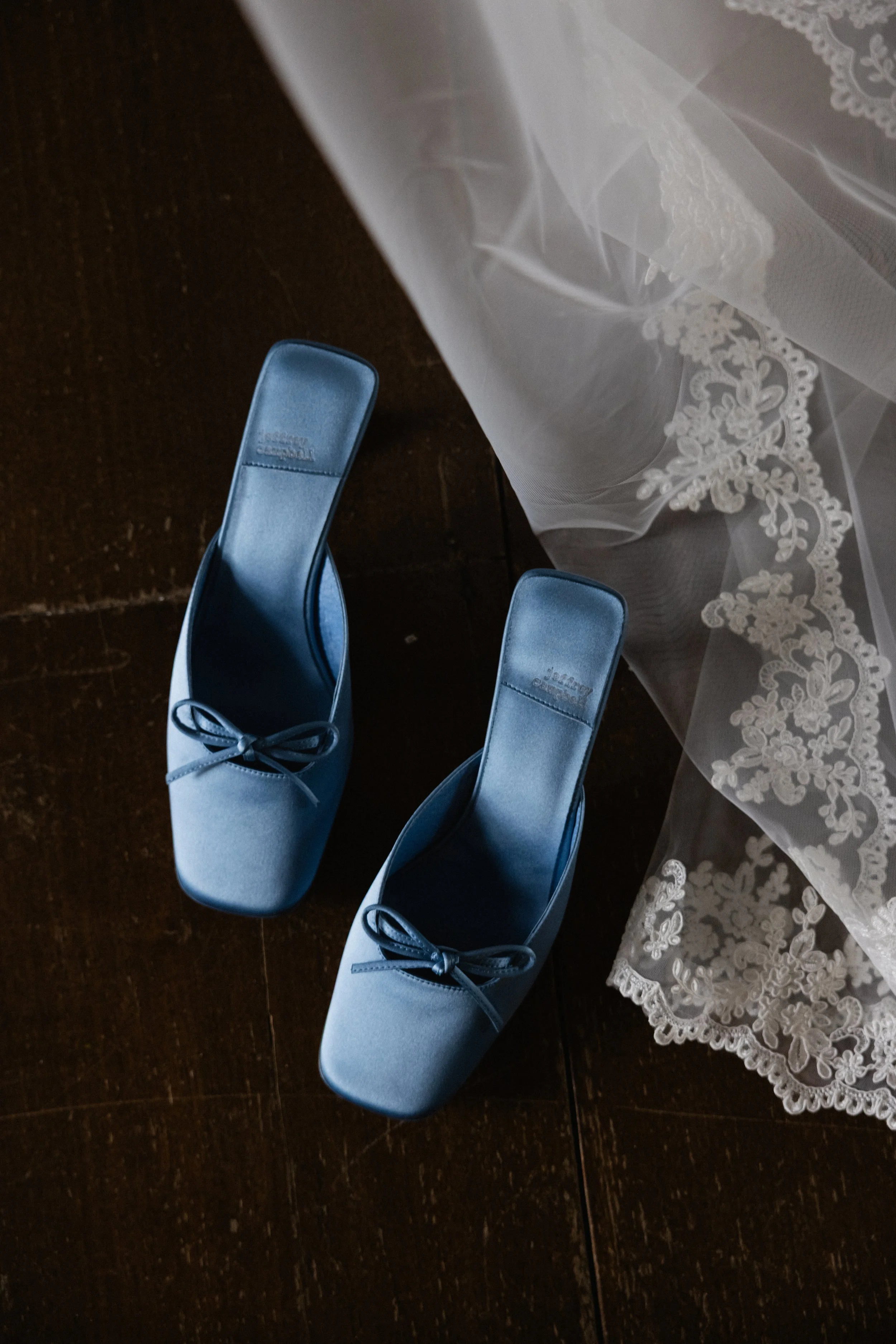 Et par blå sko med sløyfe ligger på en treparkett. Et hvitt brudeslør med blonder ligger i nærheten.