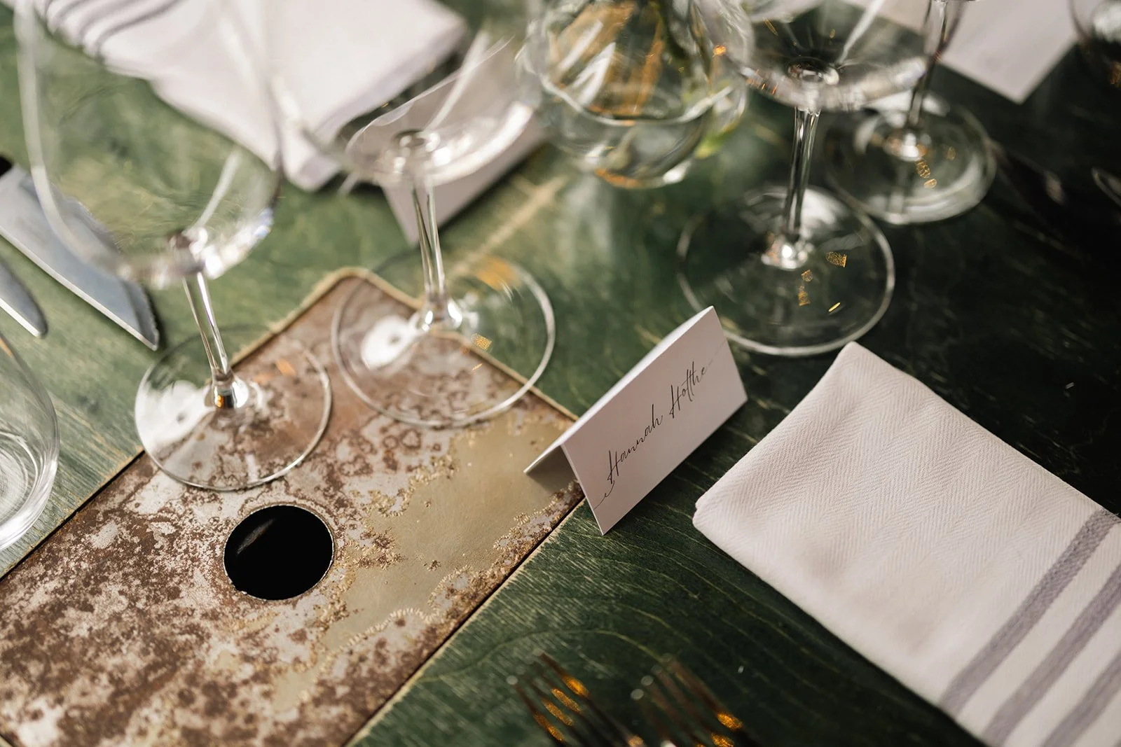 Et bord dekket til en fest eller bryllup med tomme vin- eller champagneglass, en navneskilt med navnet 'Hannah Affthe', en hvit serviett, og en rustikk, dekorert borddesign på en grønn duk.