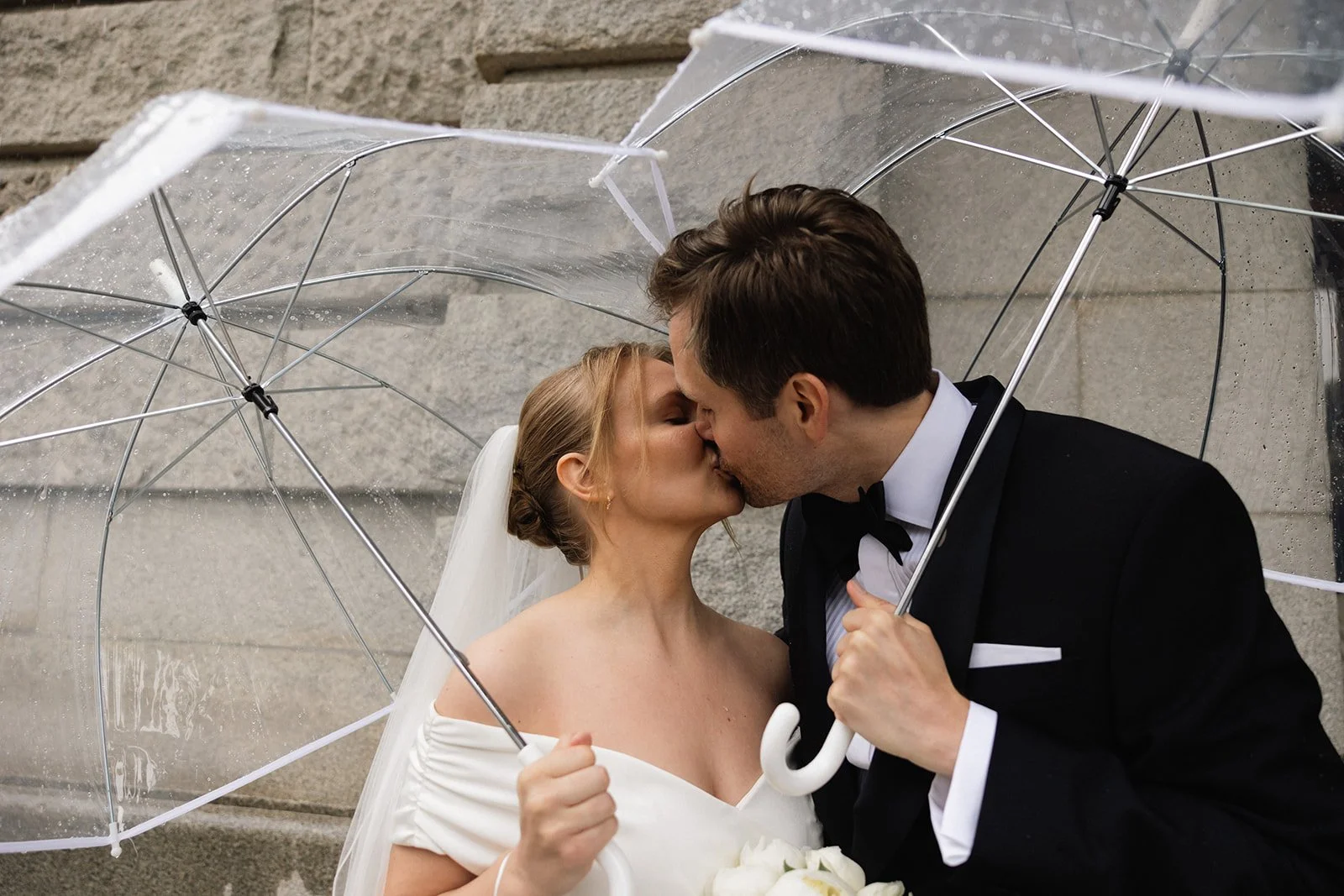 Et brudepar kysser under et transparent paraply utendørs.